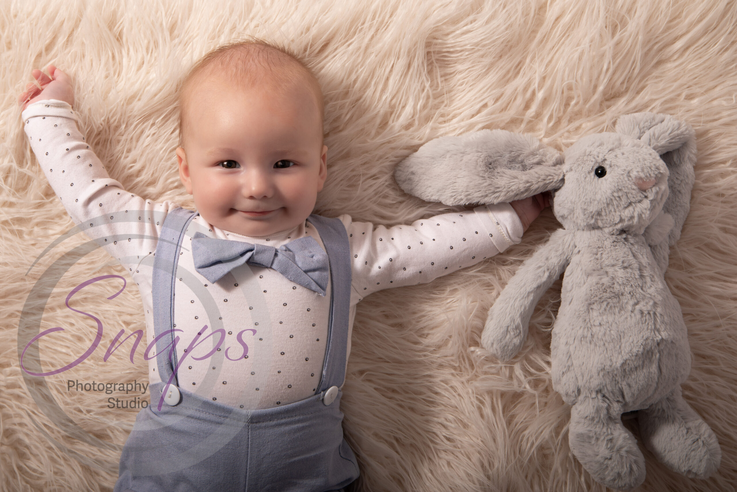 little baby boy wearing a blue bow tie and matching dungarees lead on a white fluffy rug and smiling next to his grey bunny rabbit.