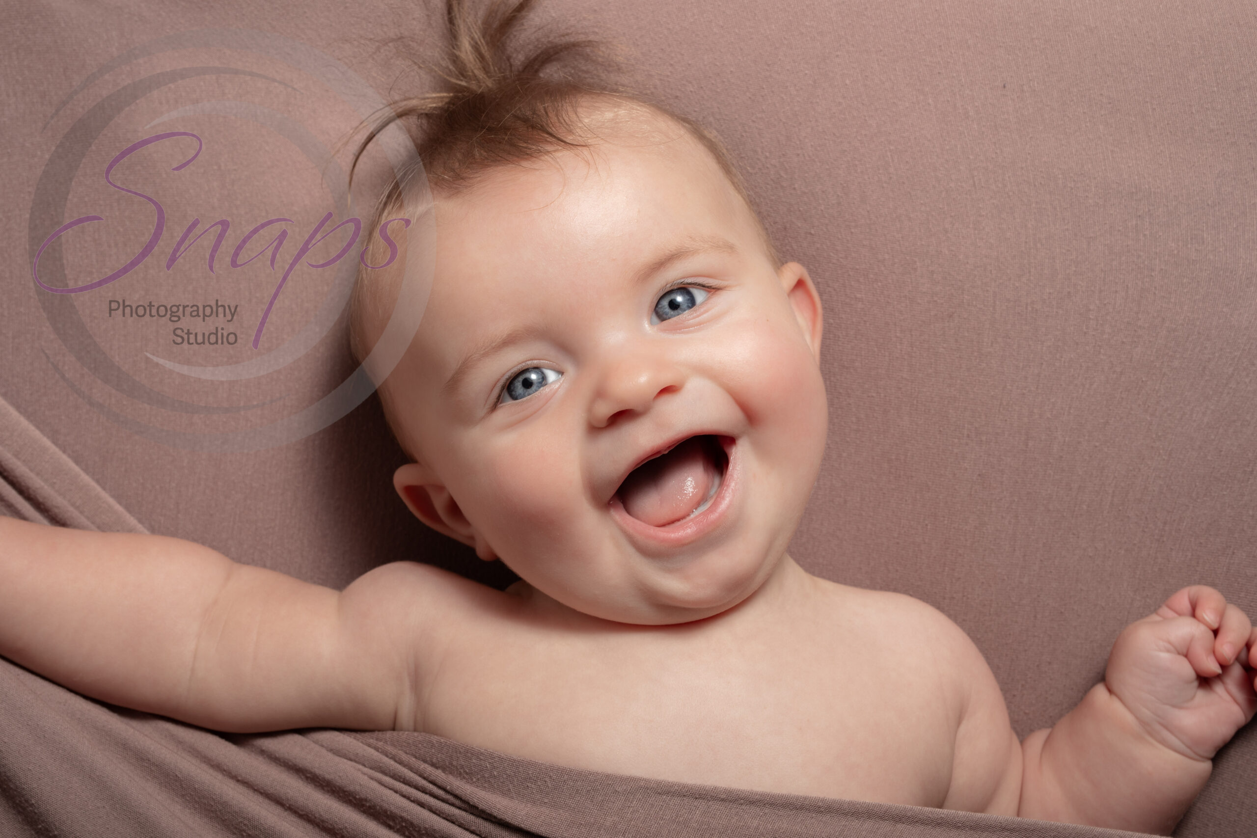 Little boy with long tuft of hair under a brown blanket laughing at the camera