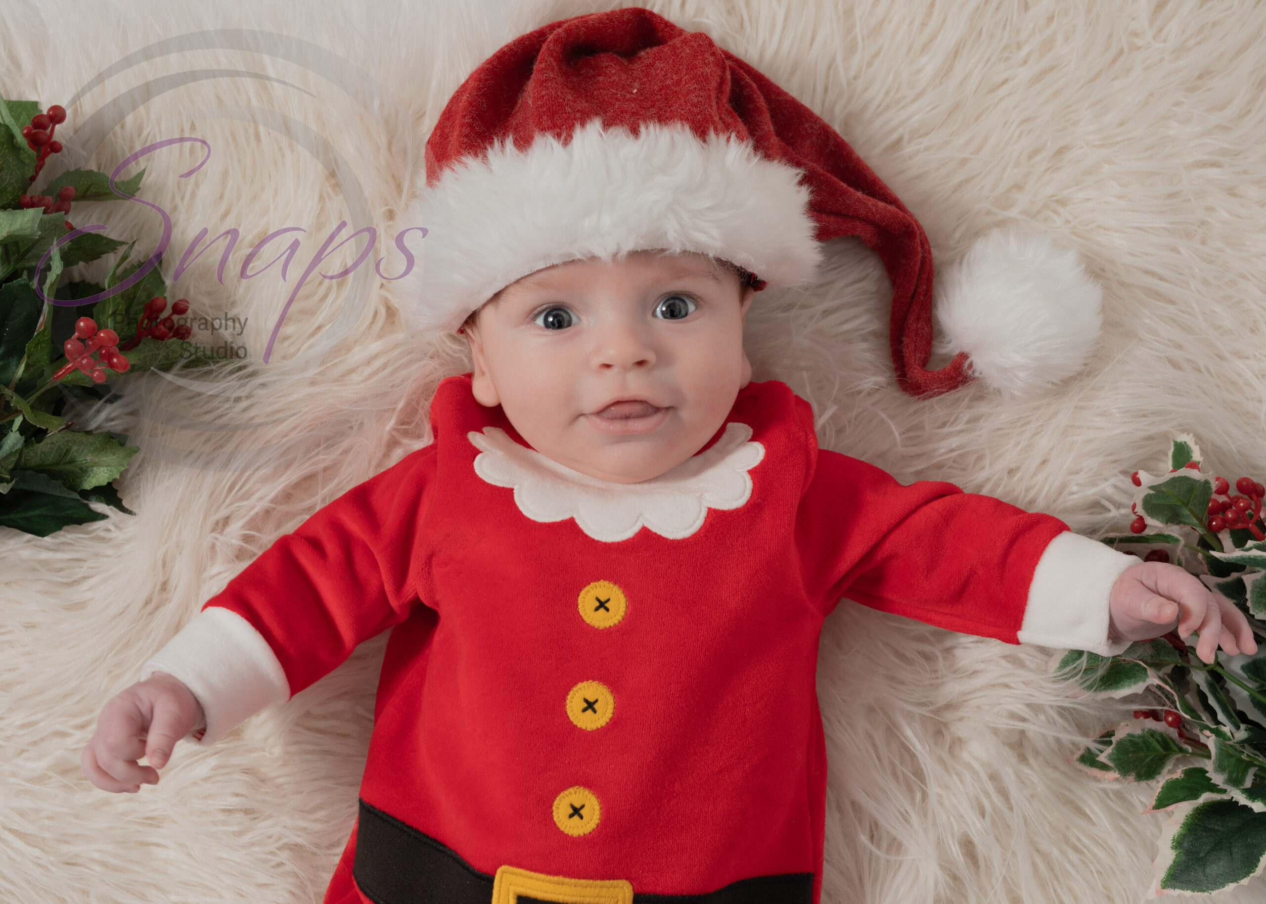baby boy with a Santa hat and Santa costume on white fluff with holly around him