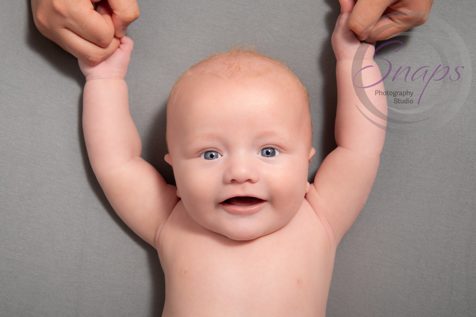 Baby boy on a green blanket smiling at the camera with his mummy holding his hands in the air.